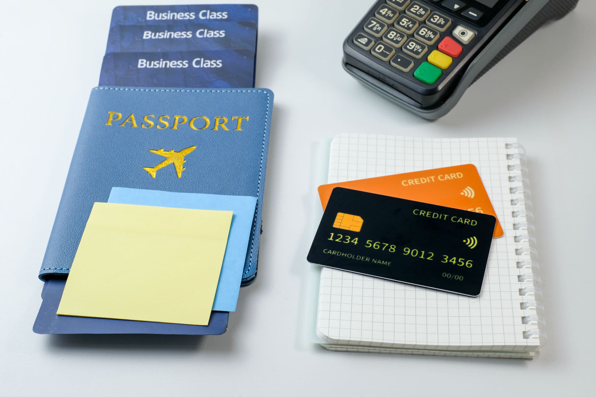 A passport and business-class tickets beside two credit cards, a notebook, and a card payment terminal on a white desk
